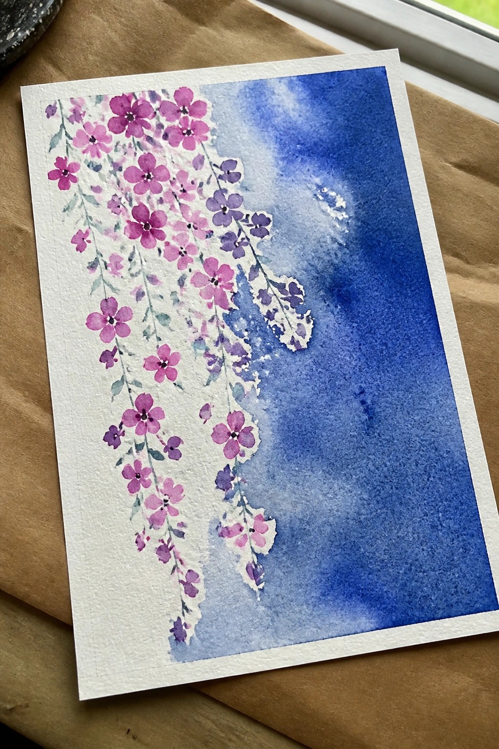 Watercolor painting of cascading purple-pink flowers beside a blue wash on white paper