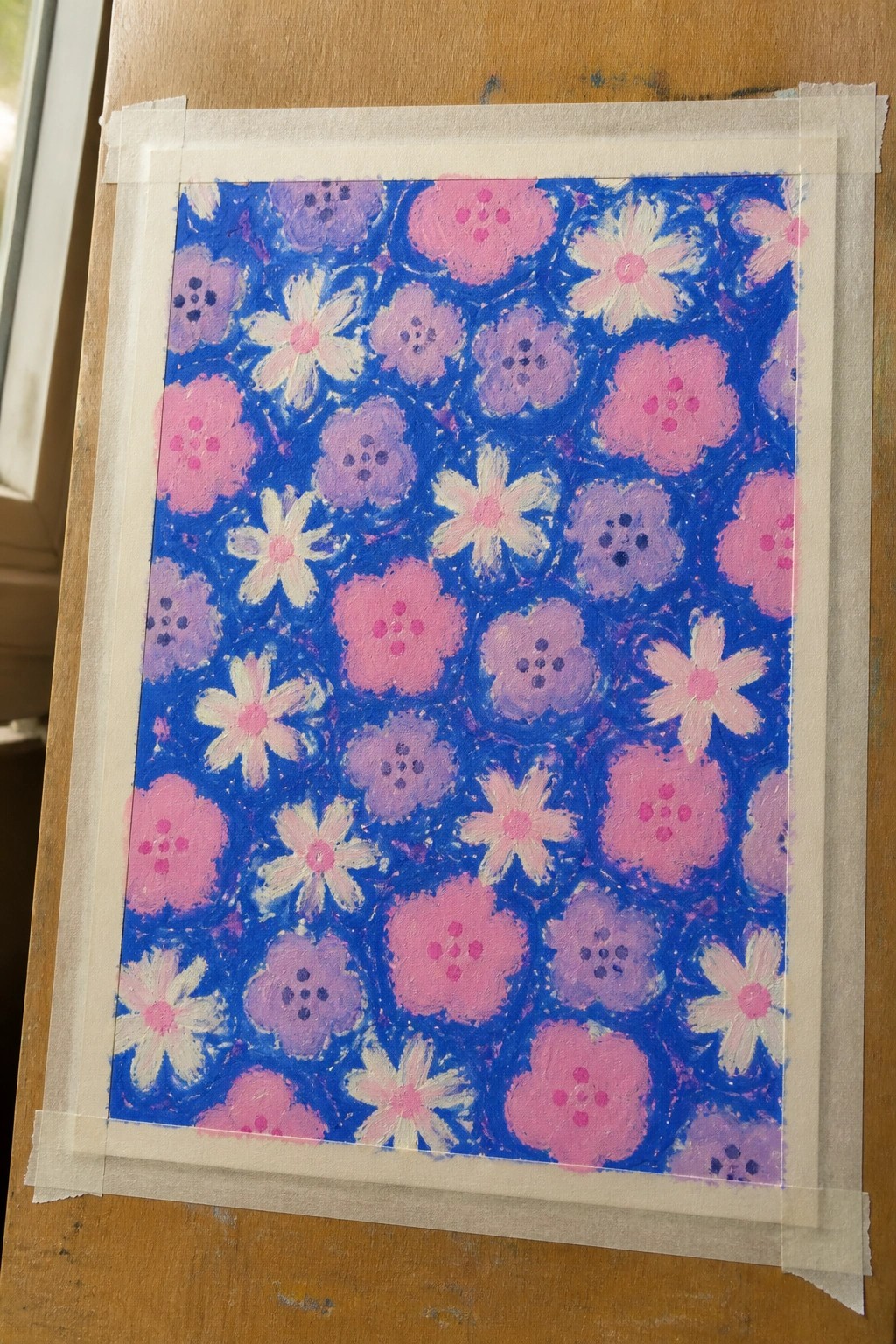 A painting of pink and purple flowers with dotted centers on a blue background, taped to a wooden surface.