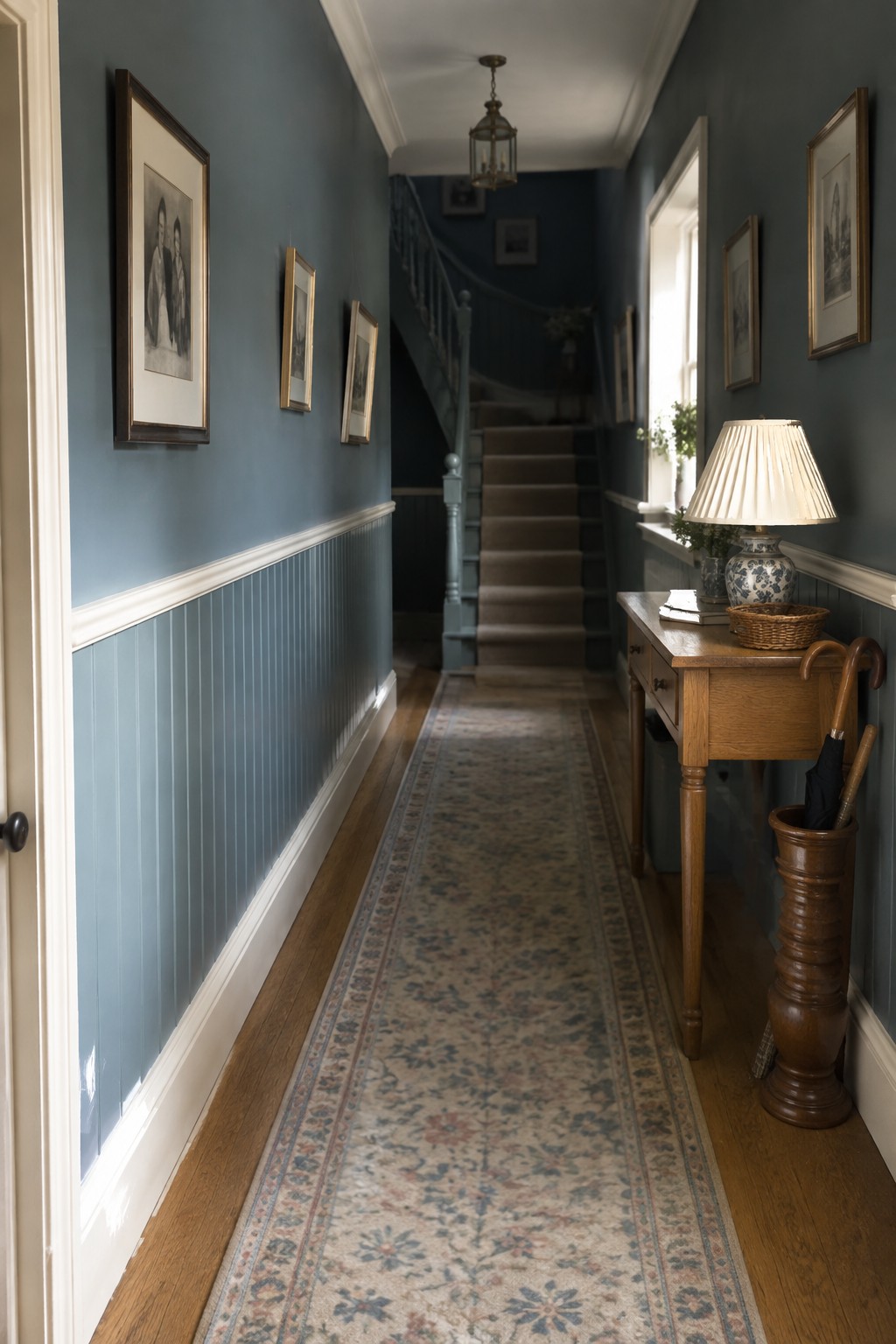 Blue hallway with framed art, wooden console table, lamp, umbrella stand, and floral runner rug.