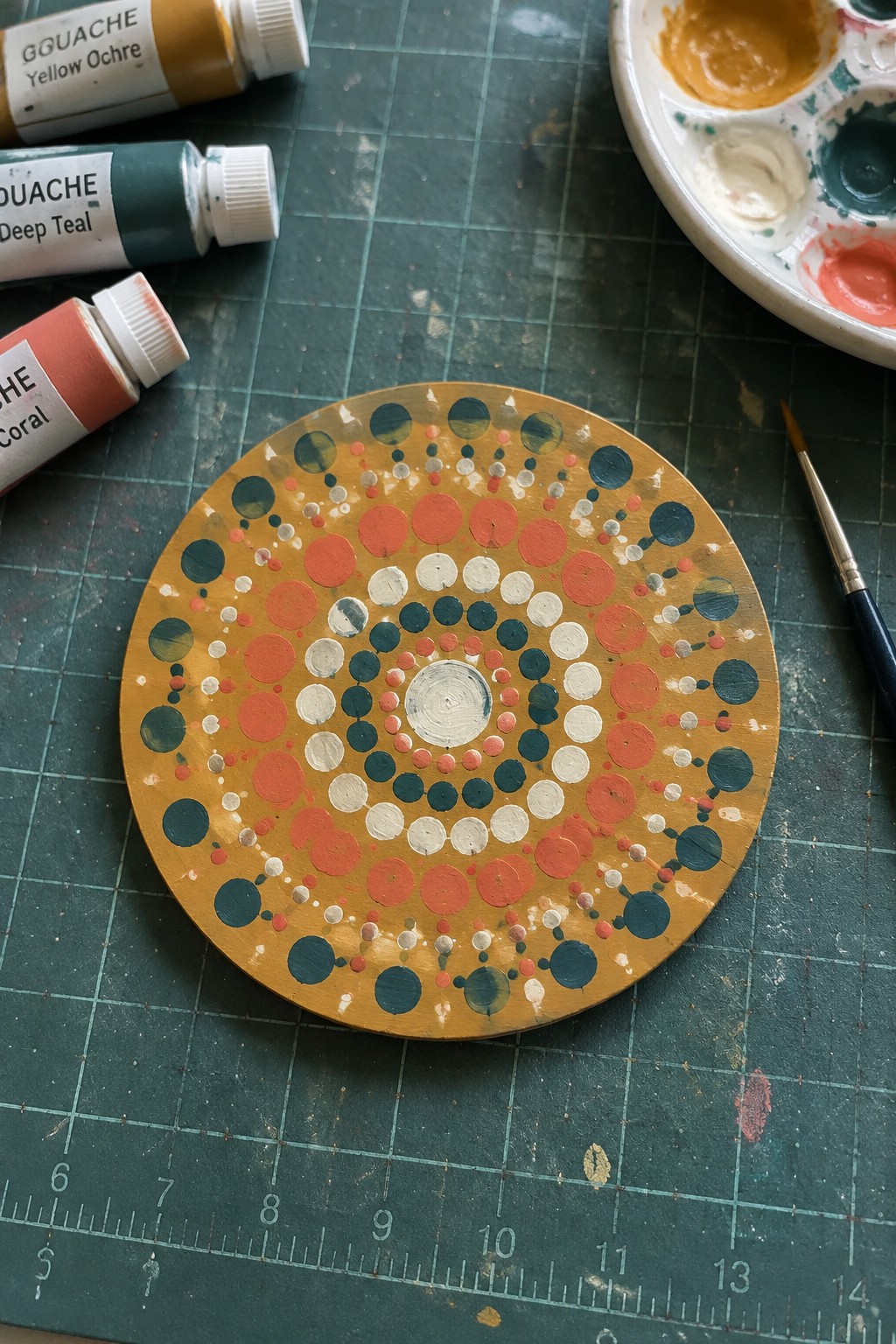 A round canvas with concentric rings of teal, coral, and white dots on a yellow ochre background sits on a green cutting mat next to gouache tubes and a paint palette.