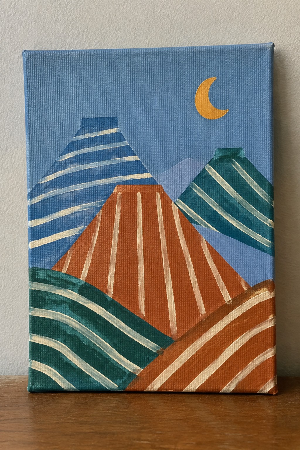 Abstract canvas painting of striped mountains in blue, orange, teal beneath yellow crescent moon