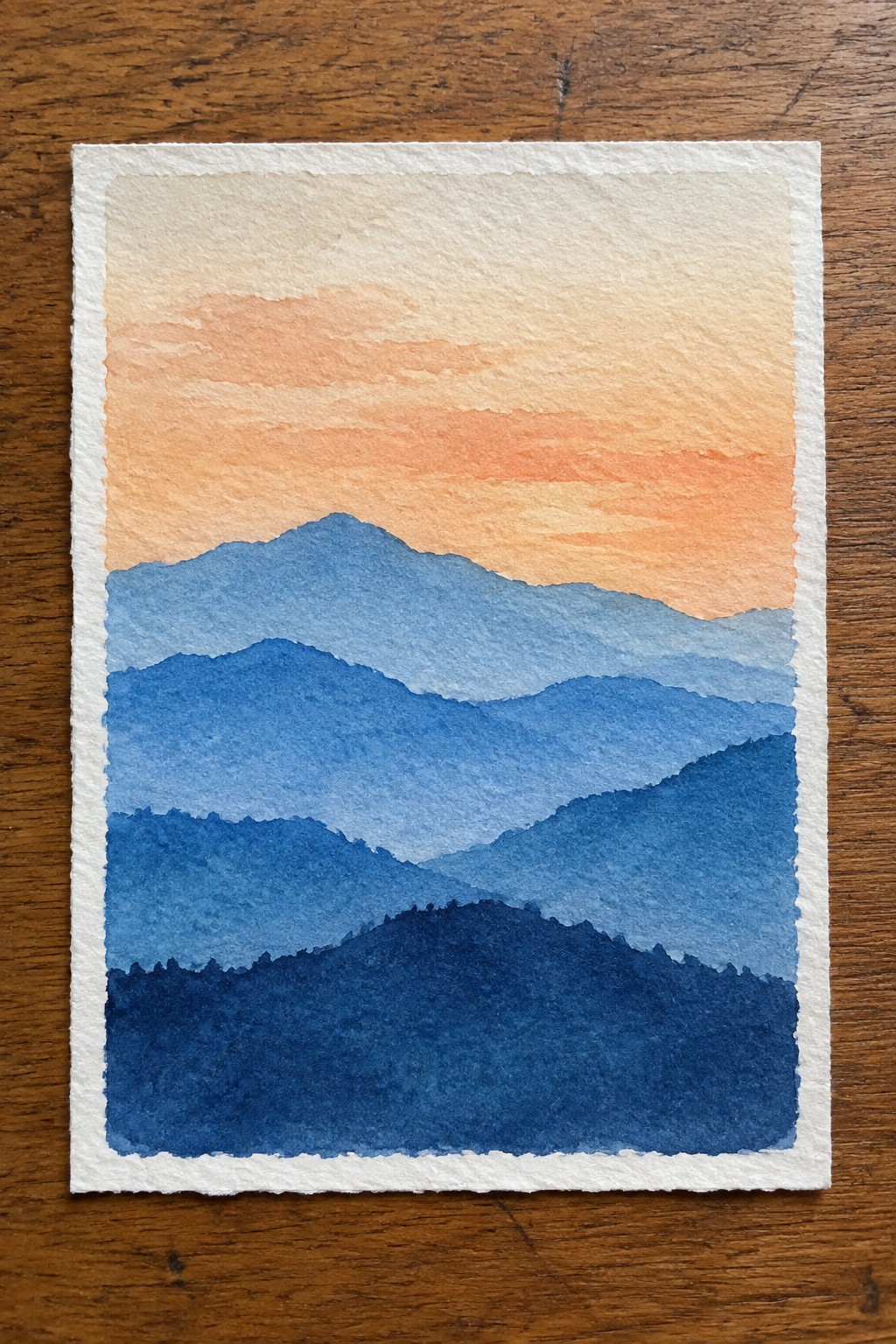 Layered blue mountains in watercolor under a warm orange sunset sky on wood.