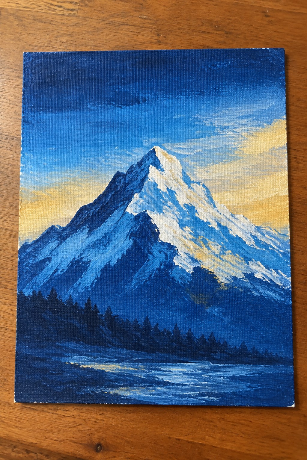 Blue-toned painting of snow-capped mountain peak with golden sky, dark trees, and water