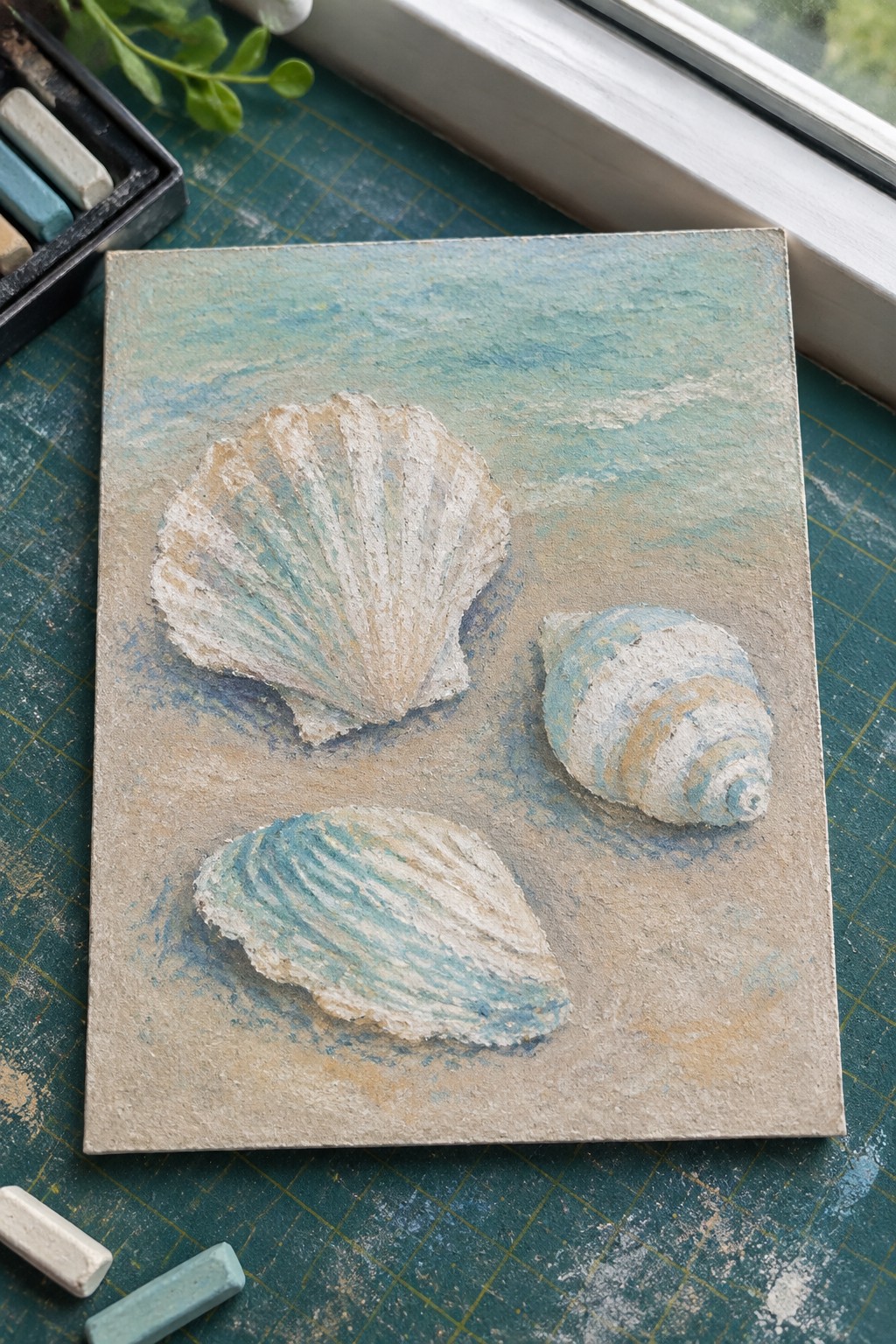 Three seashells in soft pastels on a textured blue-green background with chalks nearby