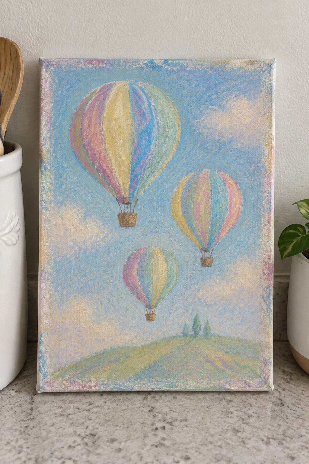 Three colorful hot air balloons float over a green hill in soft pastel painting.