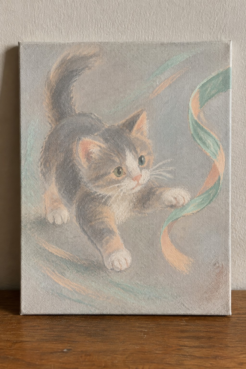 A soft pastel painting shows a calico kitten on a light background reaching out to touch a swirling green and peach ribbon.