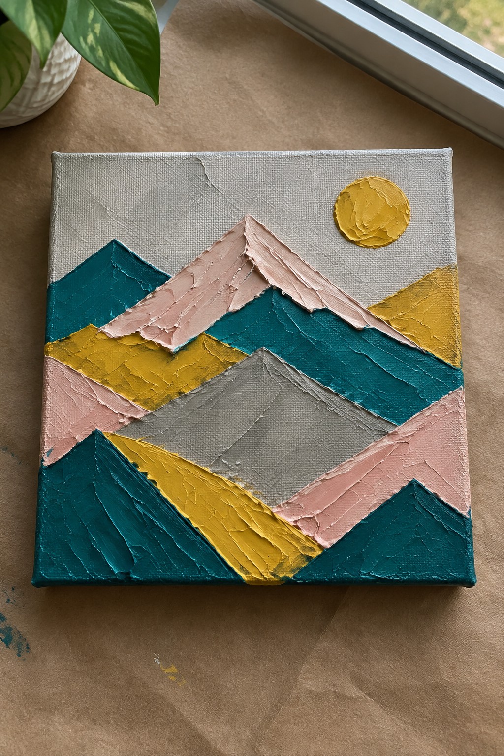 Thickly textured abstract mountain painting in teal, yellow, pink, gray with yellow sun on canvas
