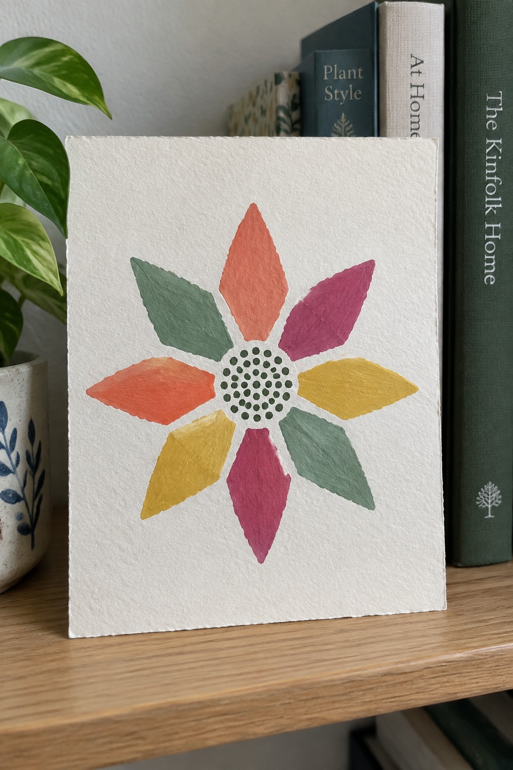 Colorful watercolor flower with eight petals on white paper beside books and plant