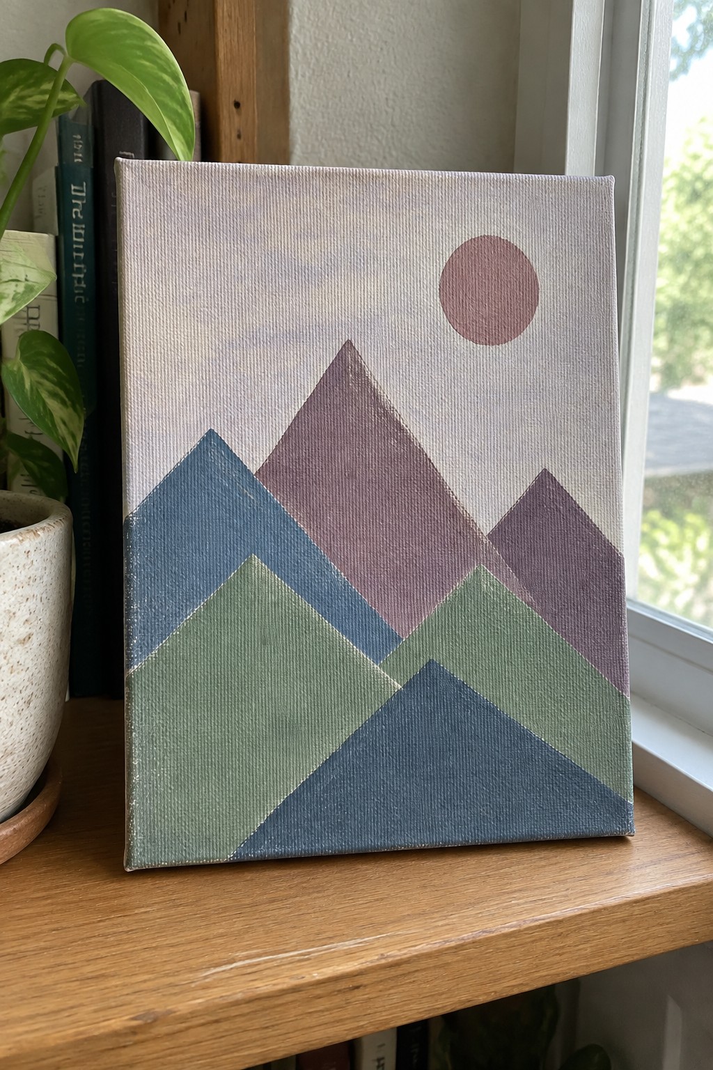 Abstract canvas painting of geometric mountains in blue green purple with pink sun on windowsill