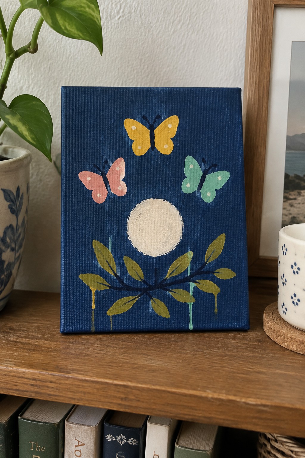 Blue canvas painting of three butterflies, white circle, and leafy branch on wooden shelf.