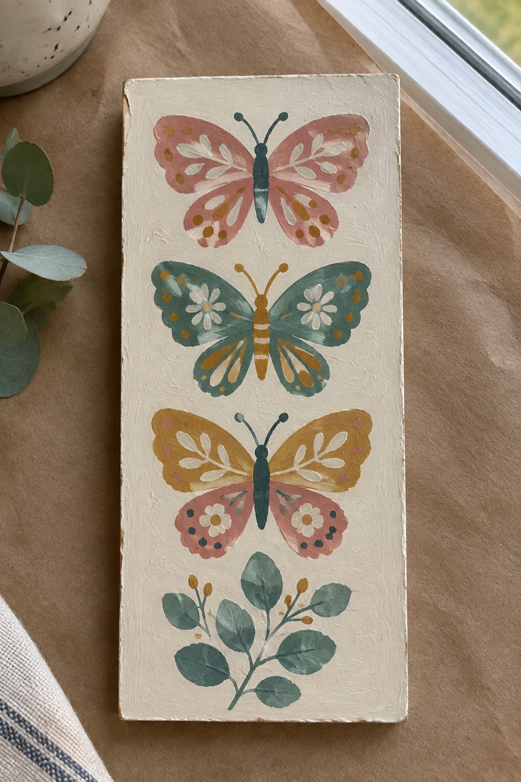 Three colorful butterflies above a green plant on a cream wooden plaque
