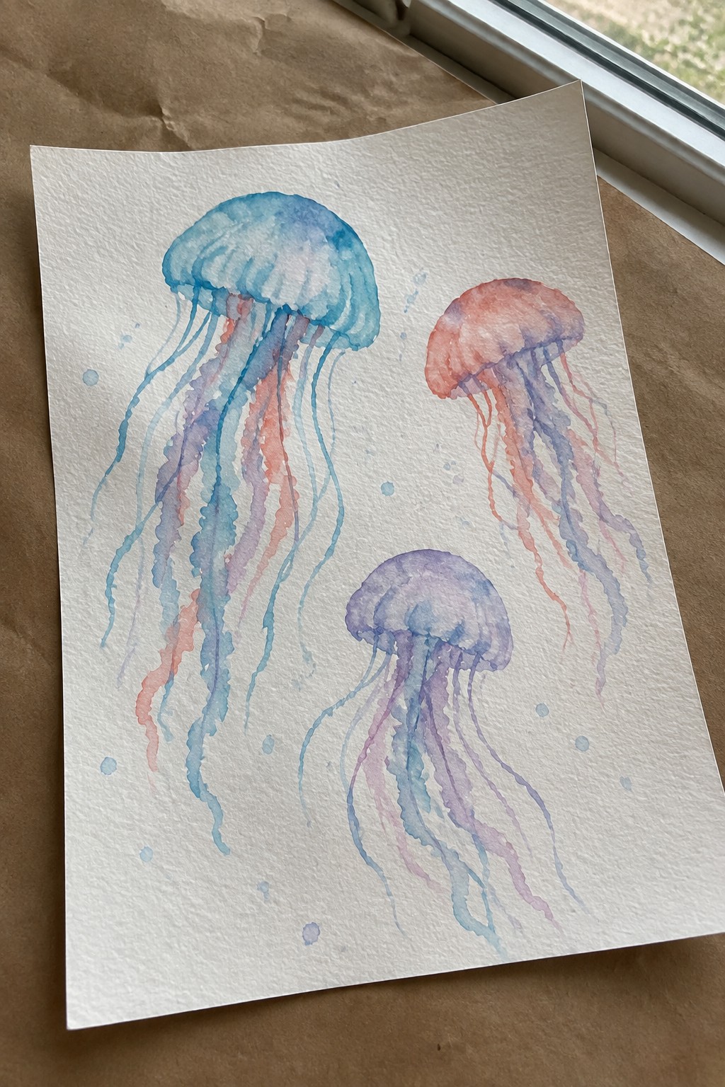 Watercolor painting of three jellyfish in blue, coral, and purple with flowing tentacles