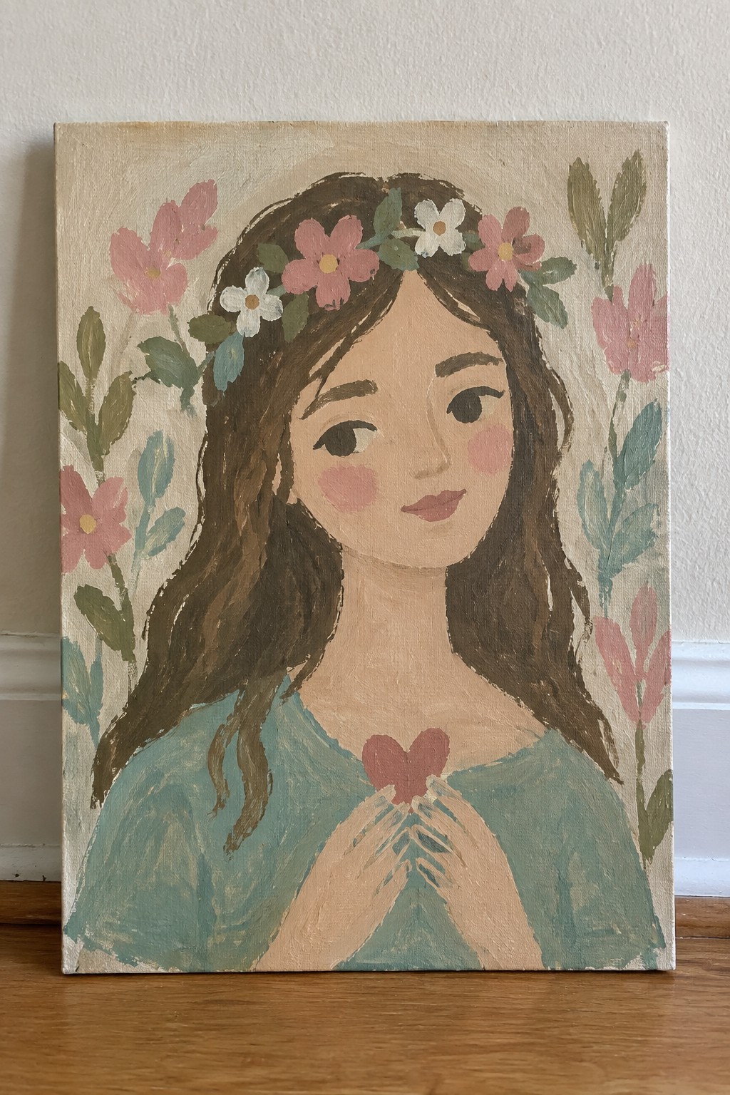 Woman with flower crown in teal top holds red heart amid pink blooms on canvas.