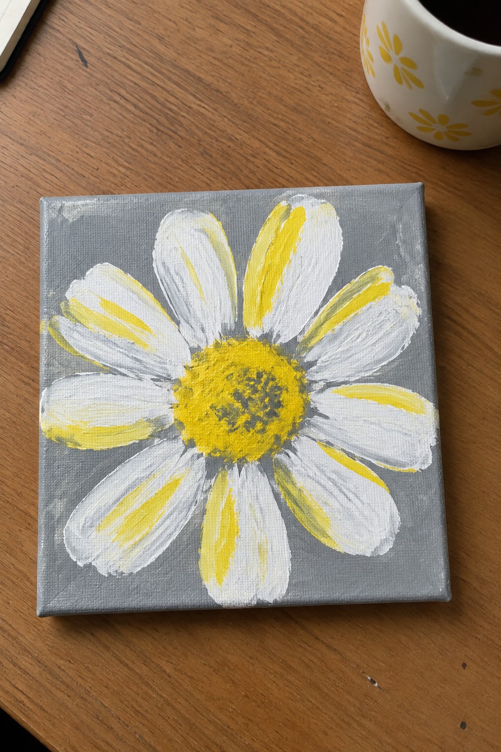 Painted white daisy with yellow center on gray canvas beside floral mug on wood