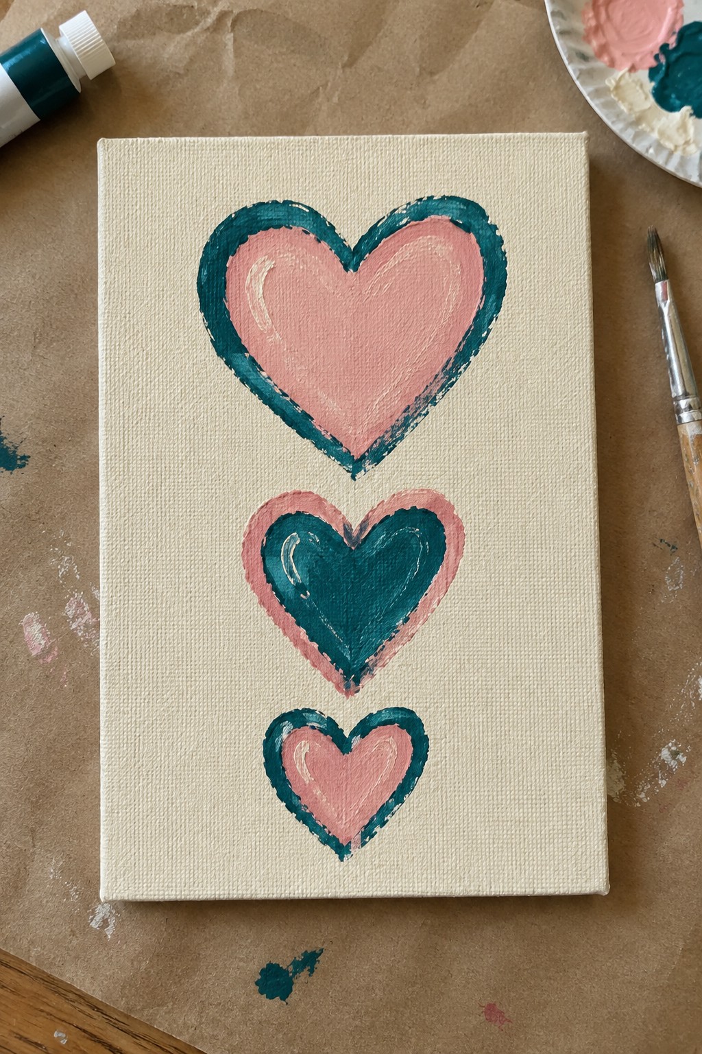 Canvas with three stacked pink and teal hearts amid paint tube, palette, and brush
