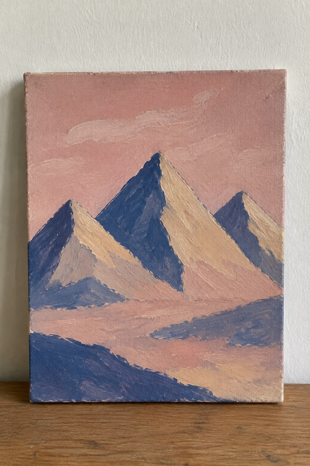 Minimalist painting of blue and beige mountains against a pink sky on a canvas.