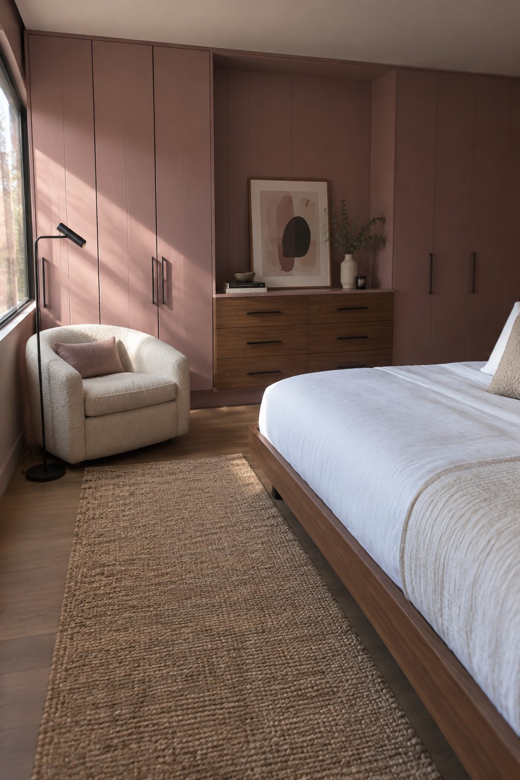 Modern bedroom with rose pink cabinetry, white bed, boucle chair, and jute rug.