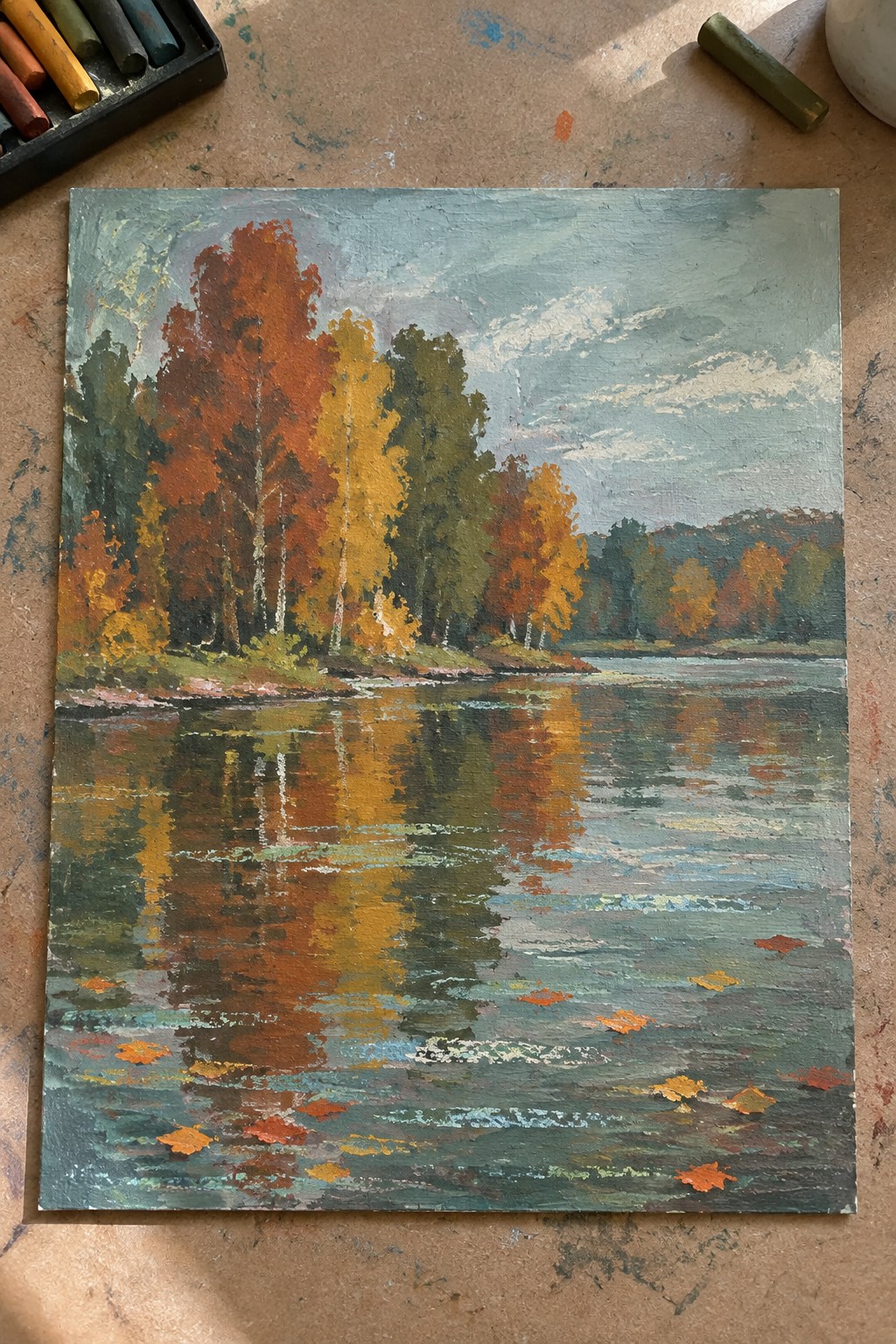 Autumn trees in red, orange, yellow reflected in calm lake with floating leaves