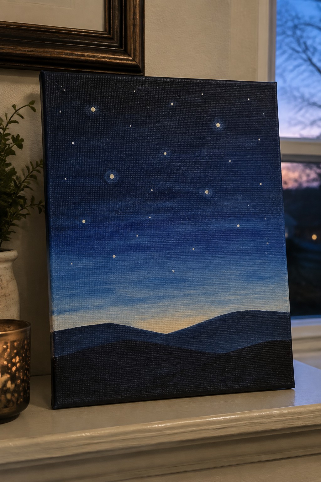 Blue gradient starry night painting with dark hills beside plant and candle