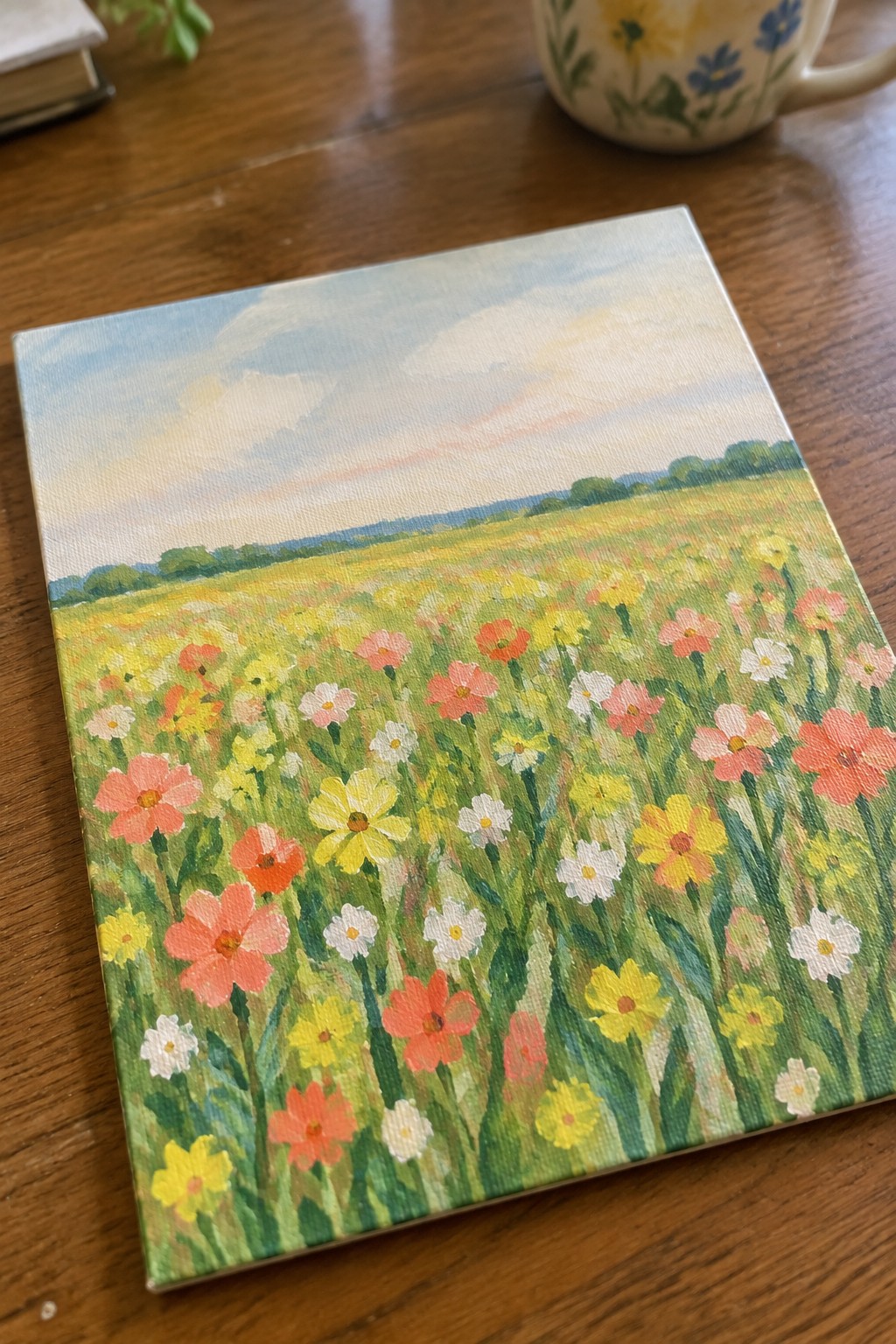 Vibrant wildflower meadow painting on canvas atop wooden table beside floral mug
