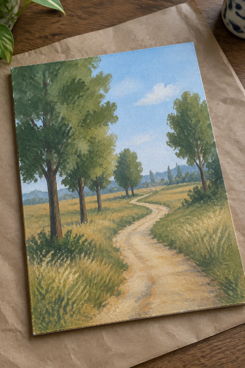 Winding dirt path through grassy fields with green trees under blue sky on canvas
