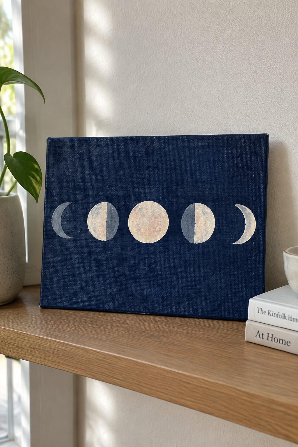 Moon phase artwork on navy canvas beside plant and books on wooden shelf