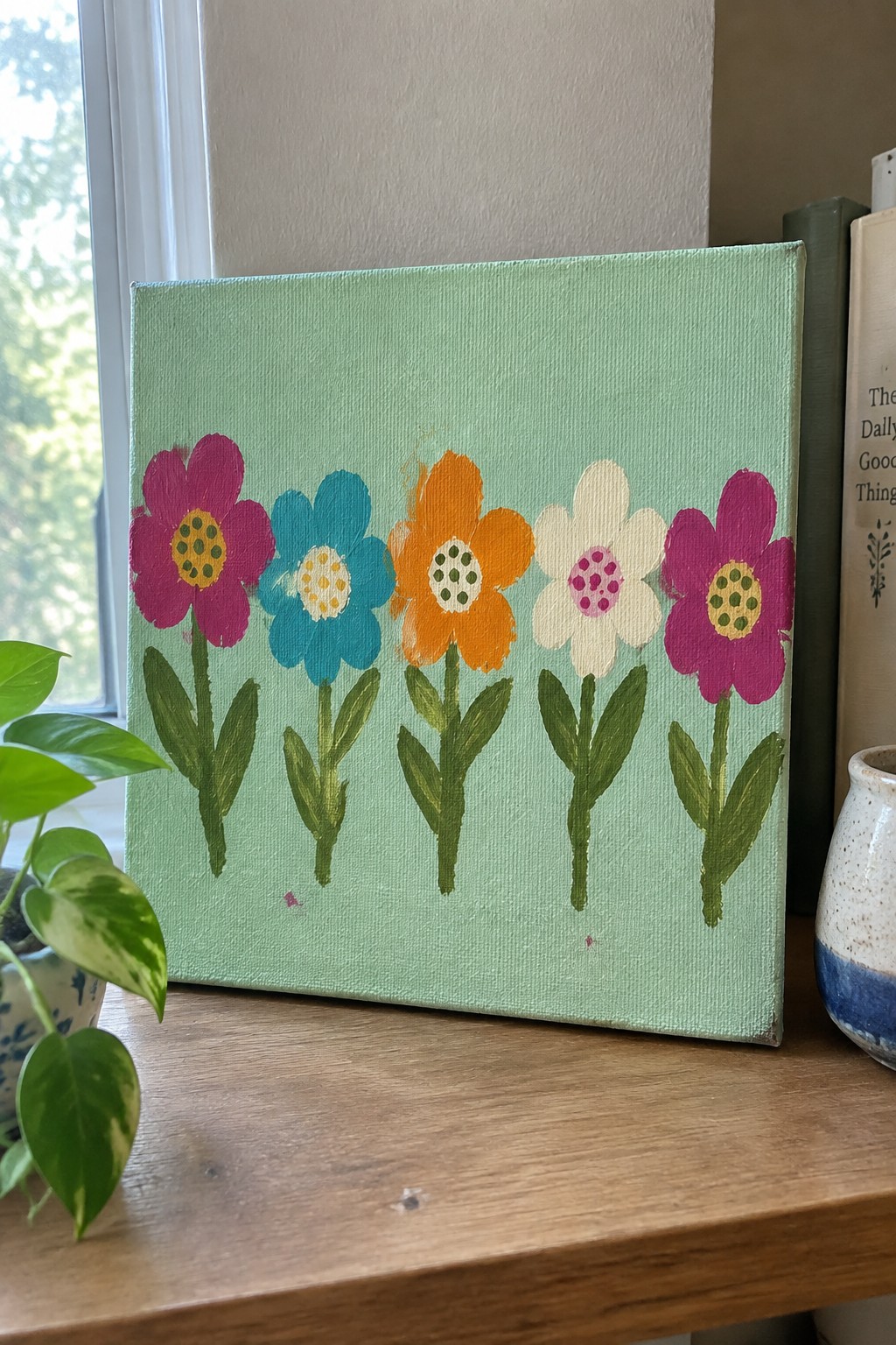 Colorful floral painting on green canvas atop wooden table with plant and books.