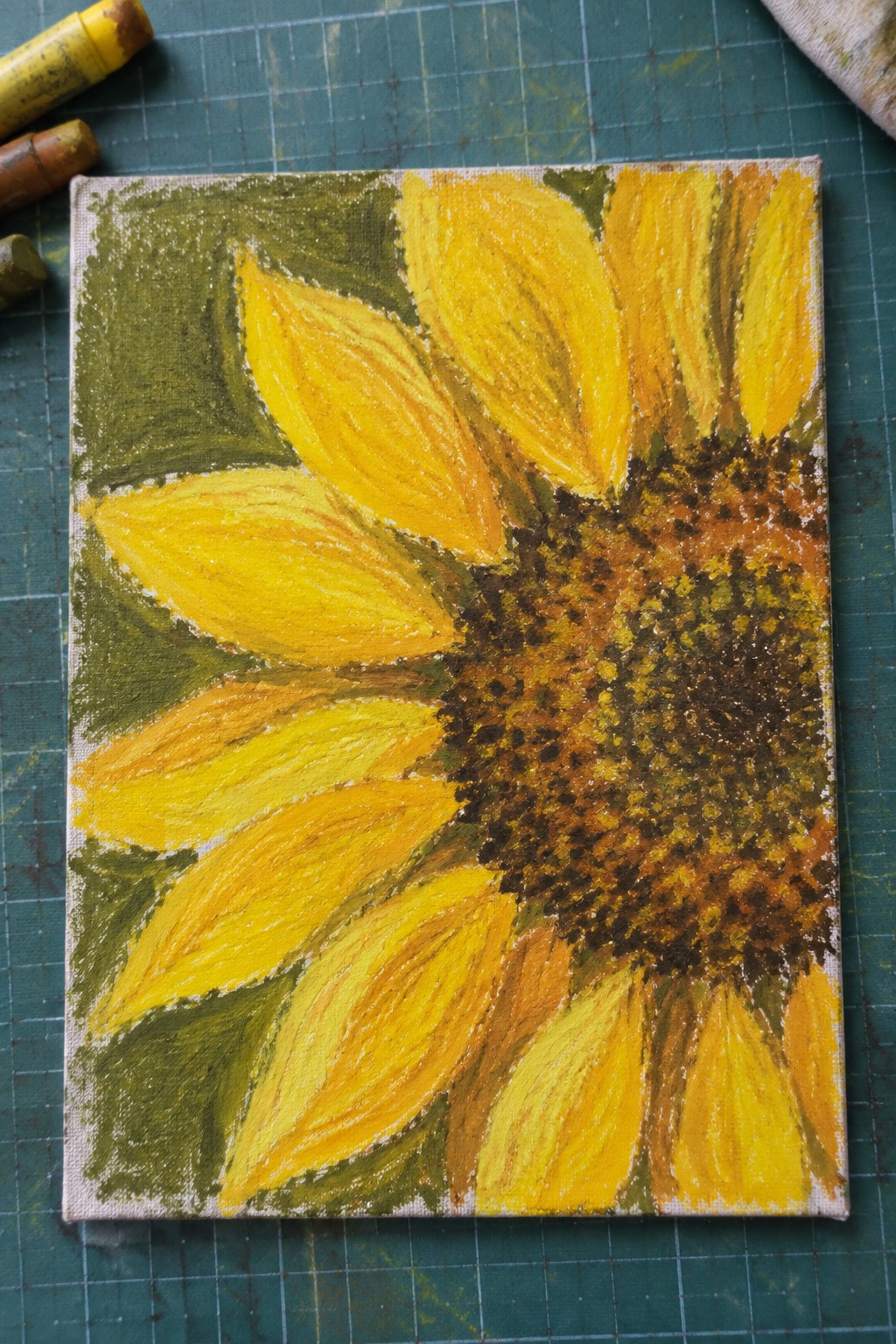 Yellow sunflower painting on canvas beside oil pastels on green cutting mat