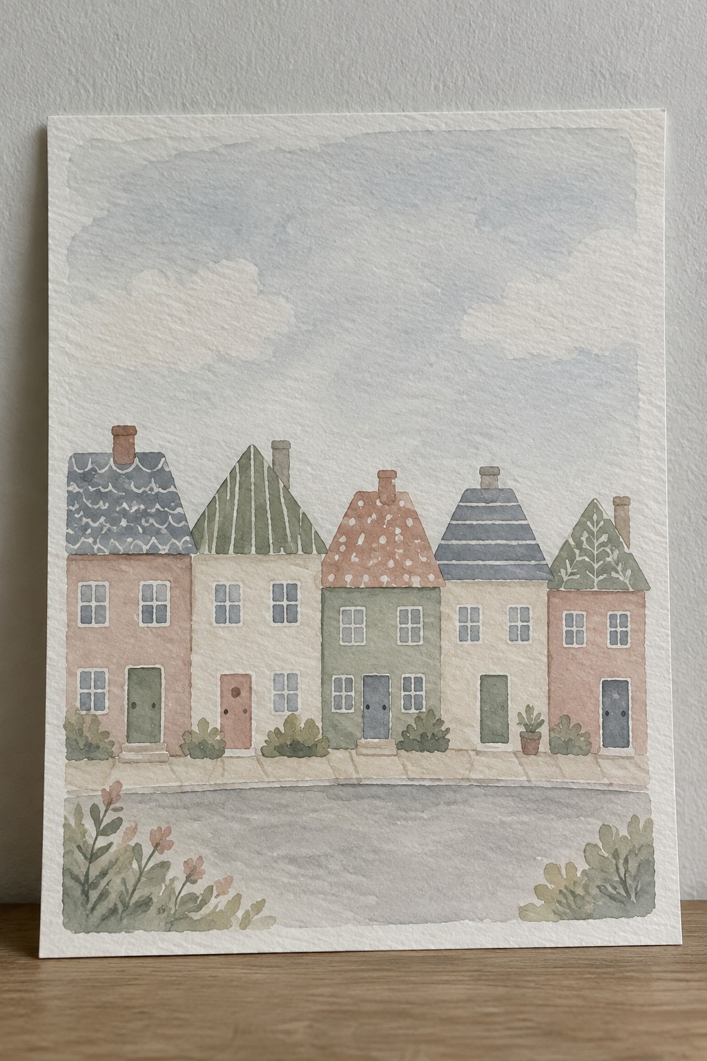 Watercolor painting of five pastel row houses with patterned roofs, chimneys, and front gardens under clouds.