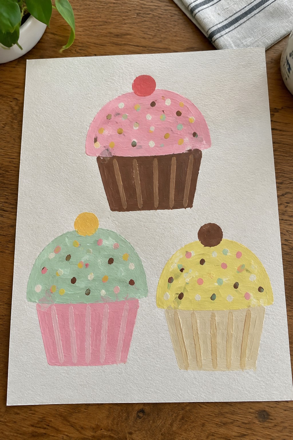 Three hand-painted cupcakes in pink, mint, and yellow with sprinkles on paper