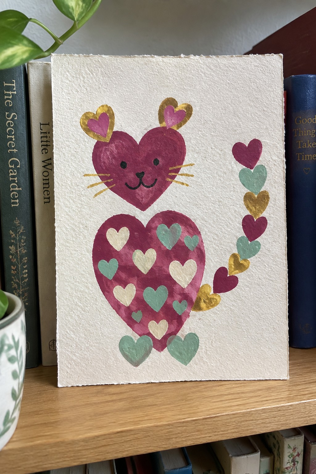 Watercolor painting of magenta heart-shaped cat with gold teal accents on bookshelf
