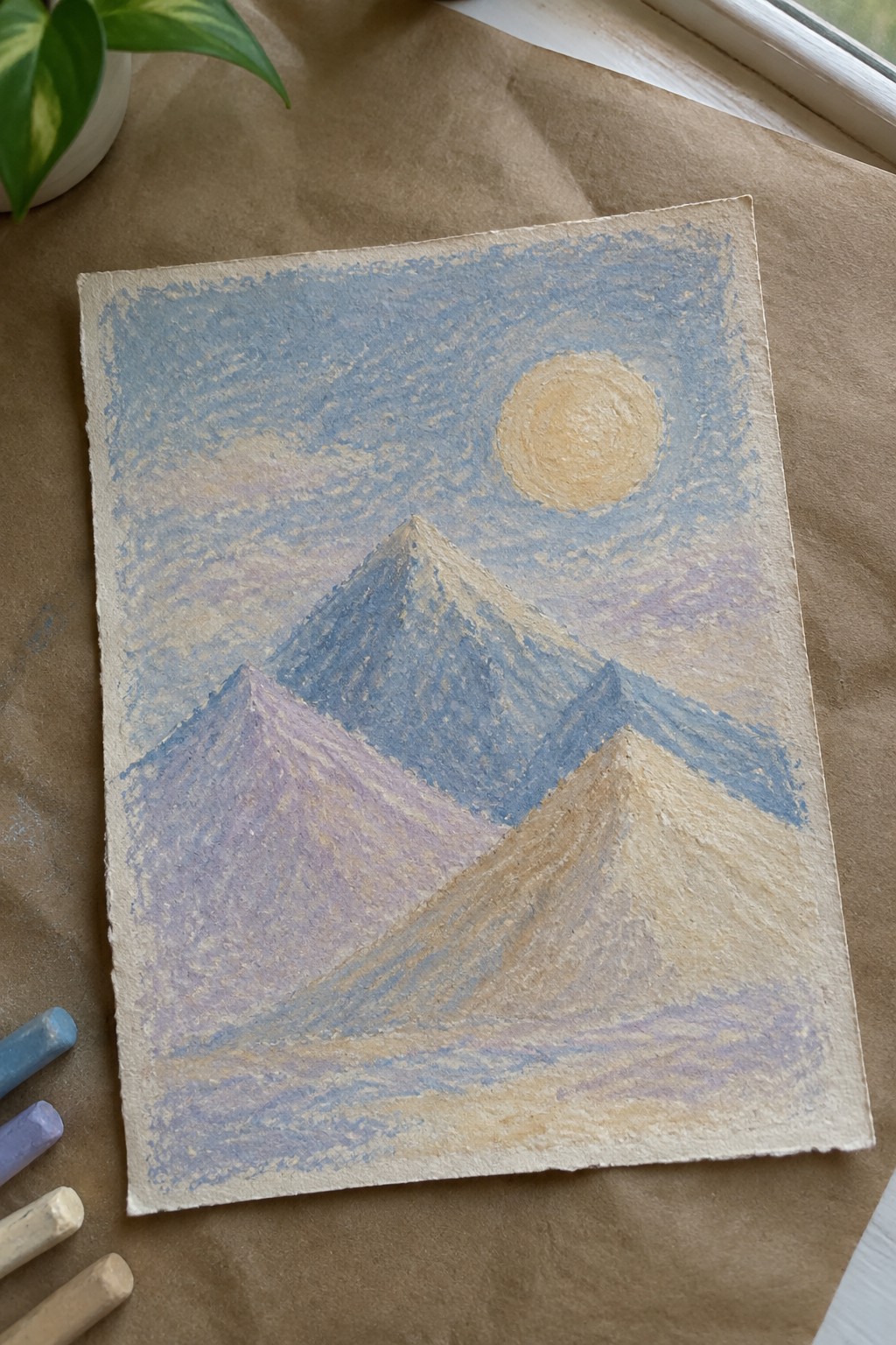 Pastel mountains in blue and purple under sun with chalks on brown paper