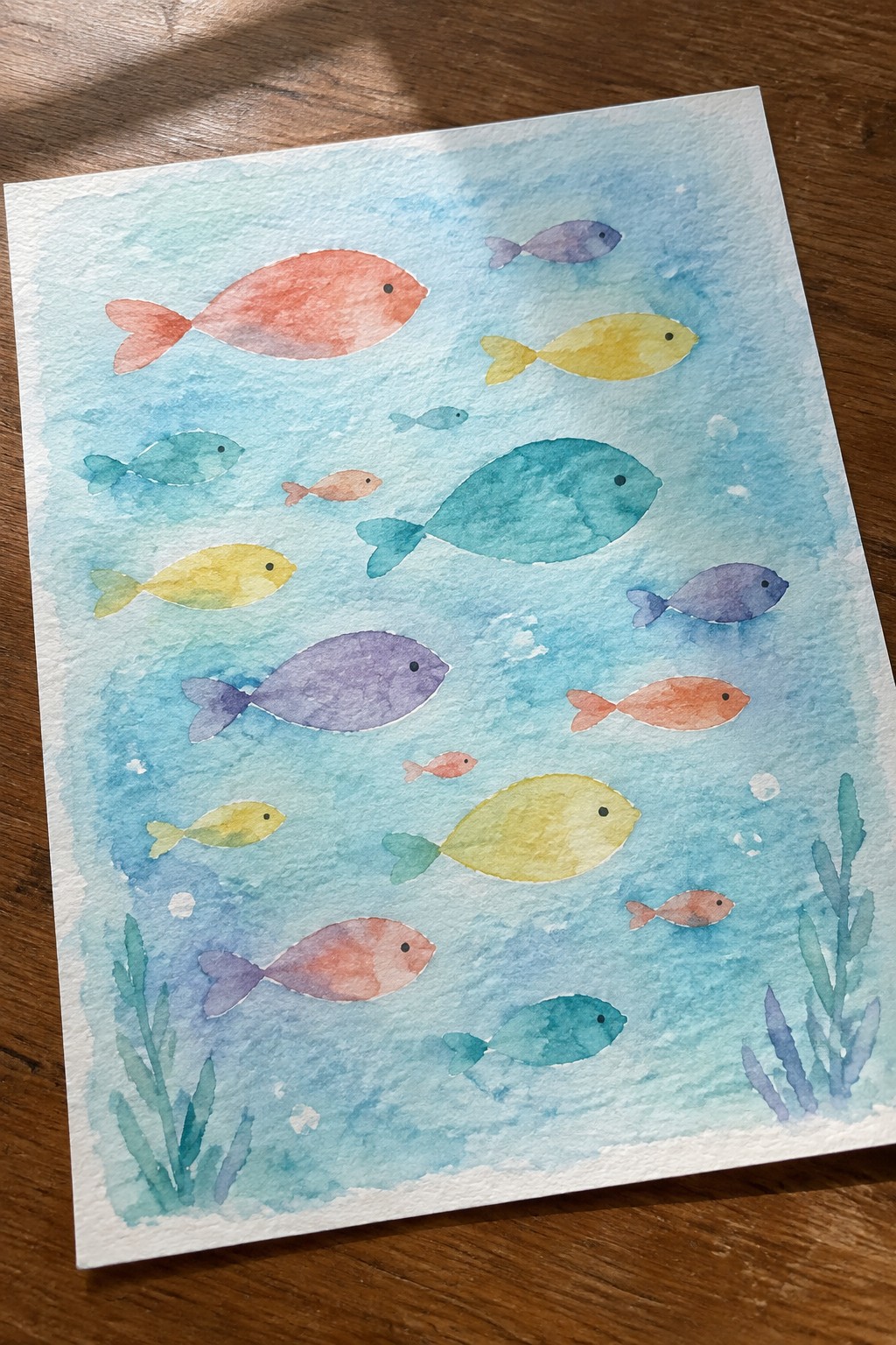 A watercolor painting of colorful fish in various sizes swimming in a light blue ocean with green seaweed at the bottom.