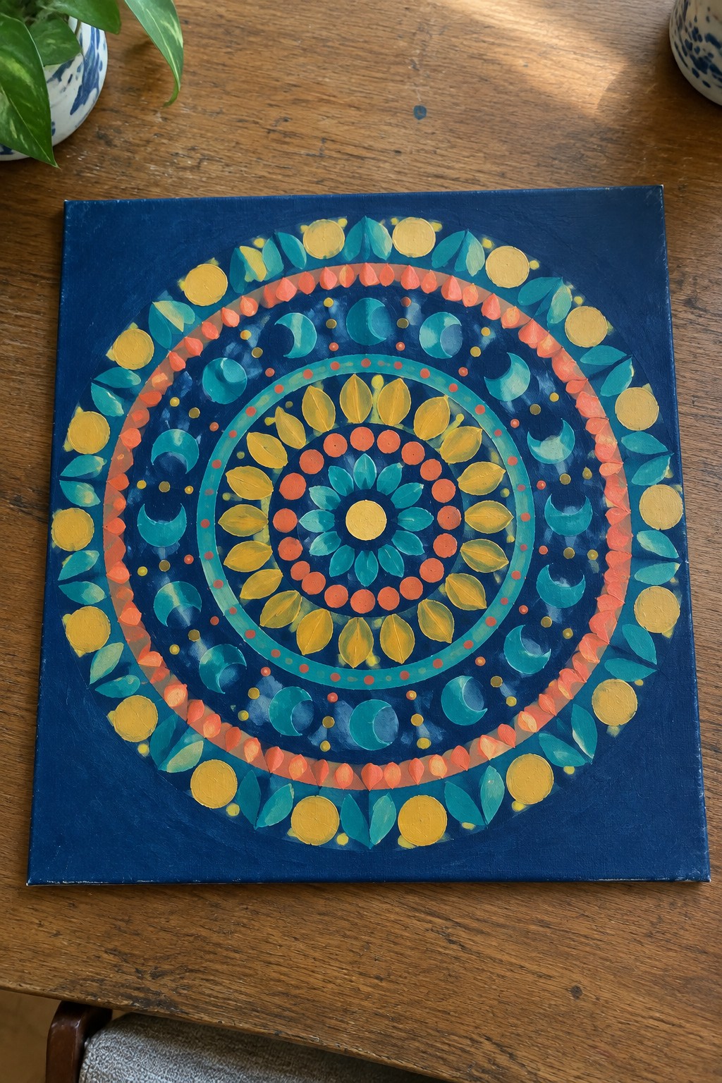 Vibrant mandala on dark blue canvas with teal, yellow, orange moons, leaves and dots on wood table