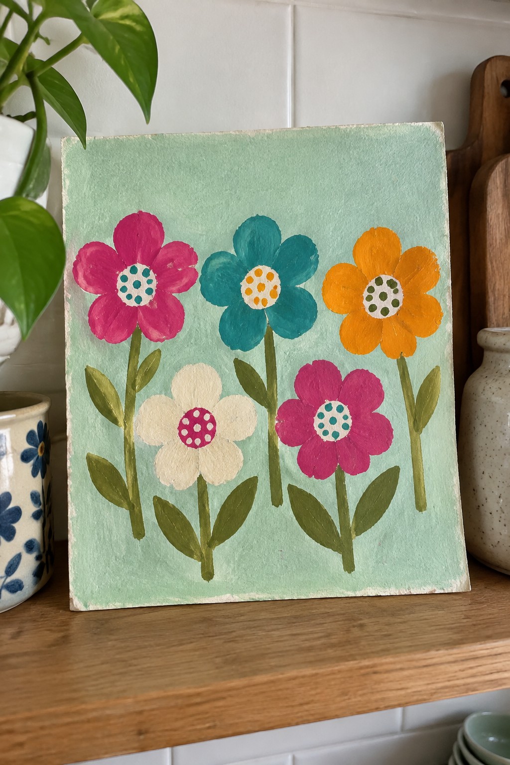 Hand-painted canvas with five colorful flowers on light green background on wooden shelf.