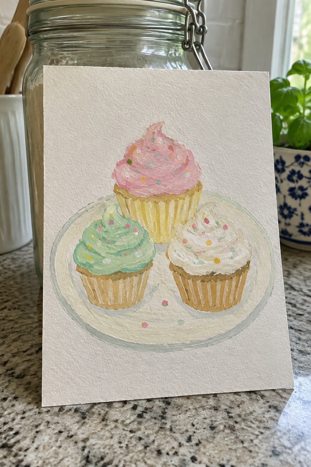 A painting of three cupcakes with pink, green, and white frosting and colorful sprinkles arranged on a round plate.