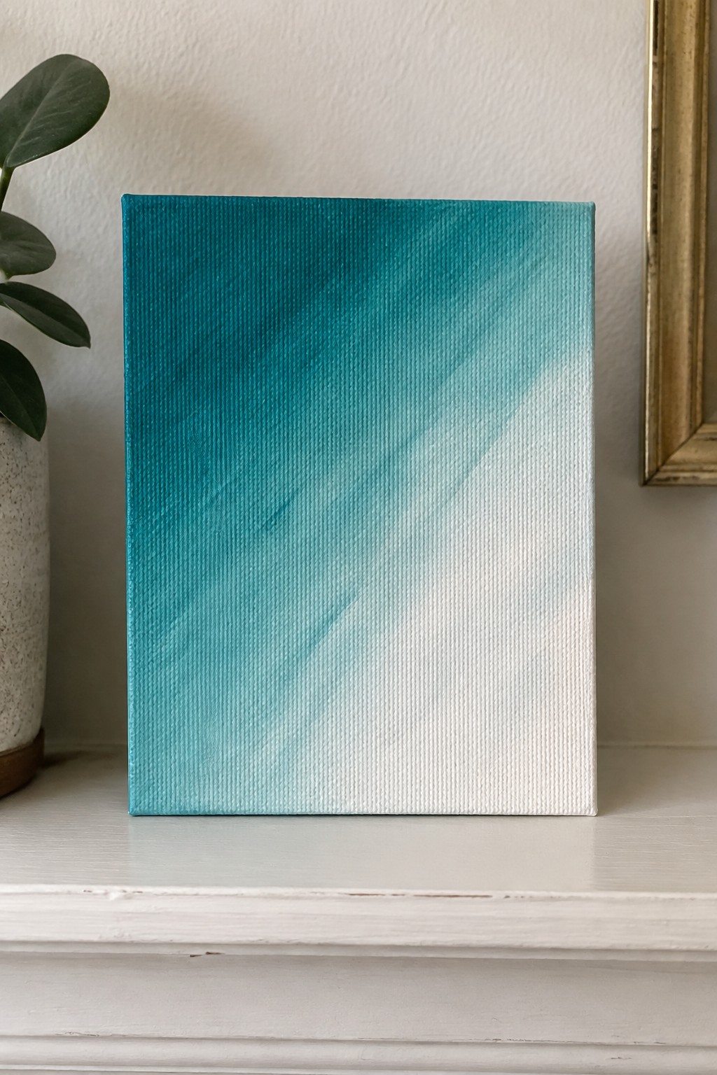 Teal gradient canvas painting on white mantel beside potted green plant