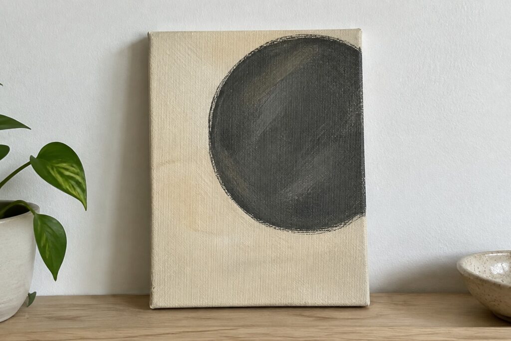 Abstract canvas with large dark gray circle on wooden shelf beside green plant