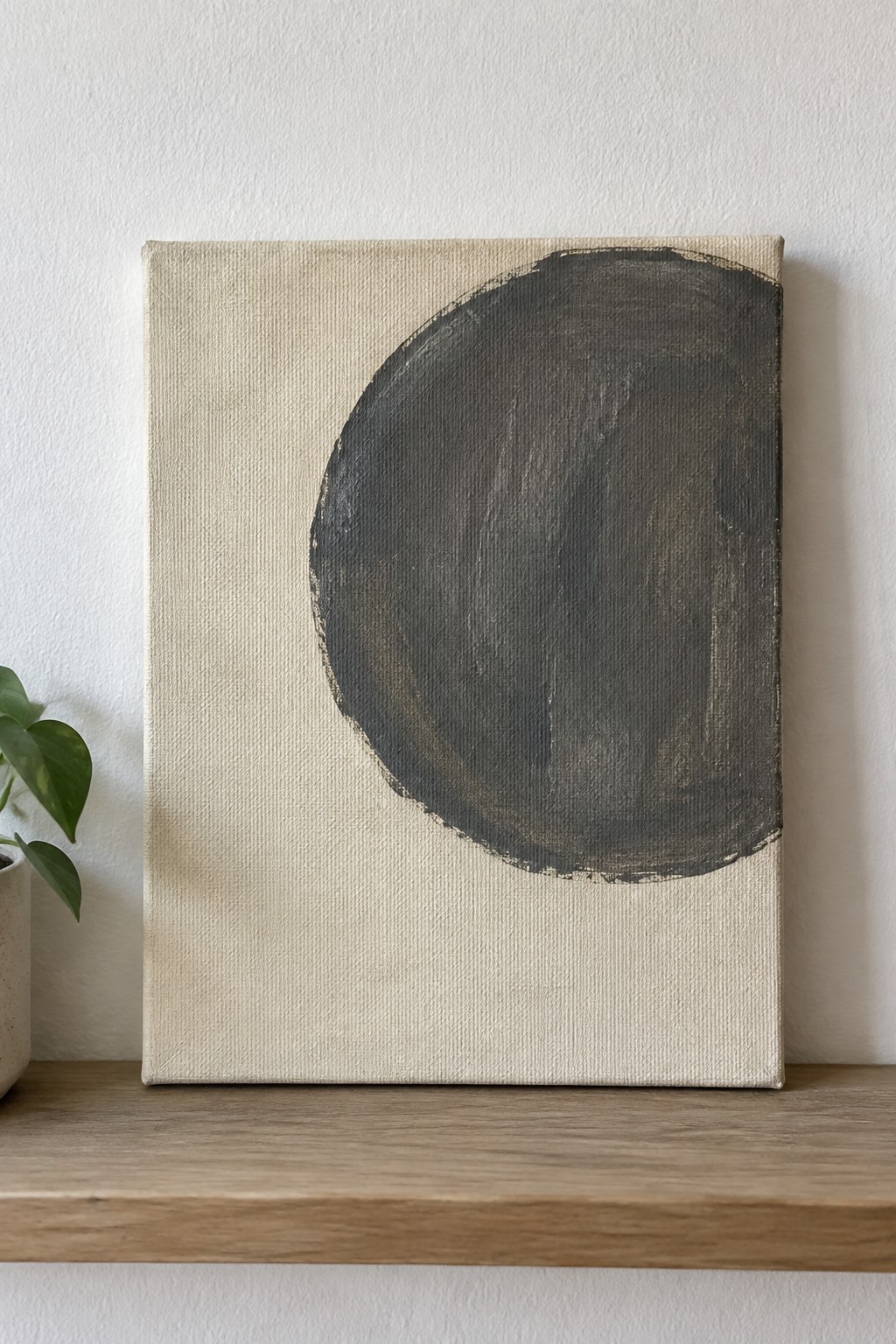 Abstract canvas with large dark gray circle on wooden shelf beside green plant