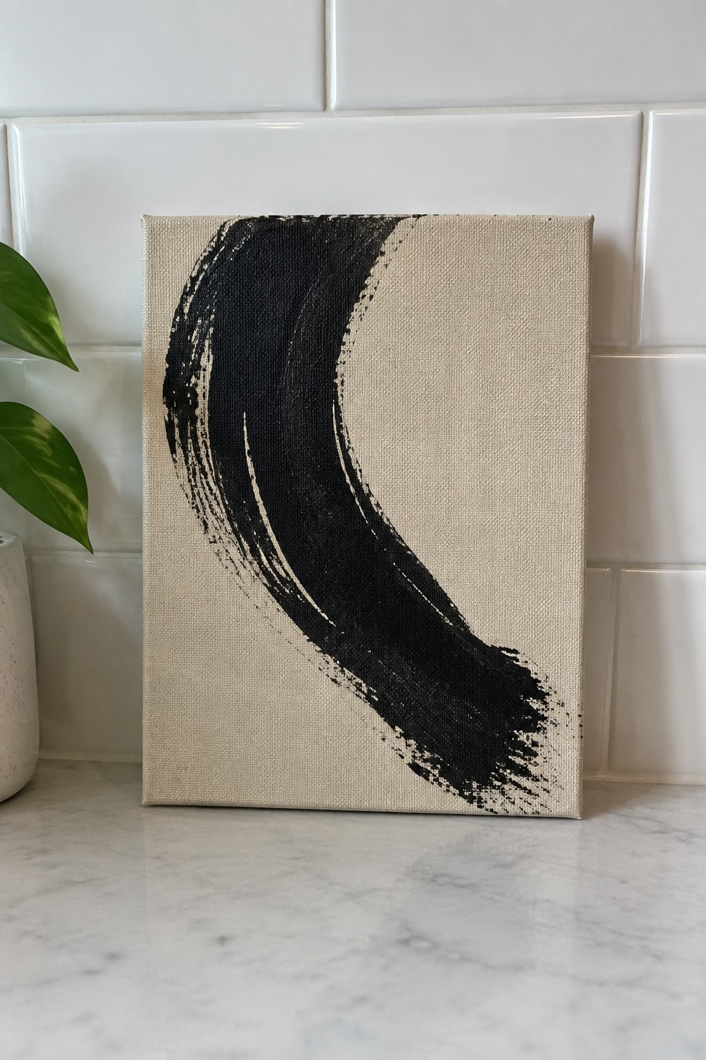 Abstract black curved brushstroke on beige canvas atop marble counter near green plant