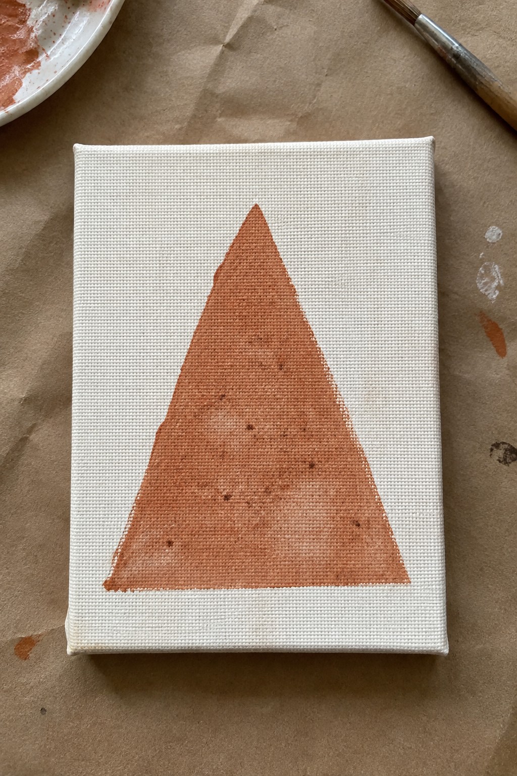 Mini canvas with centered orange triangle on brown paper near paintbrush and palette