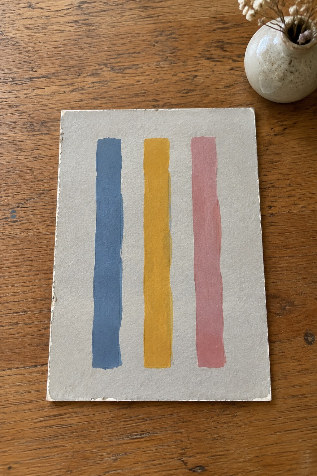 Three vertical stripes in blue, yellow, and pink on textured paper beside small vase