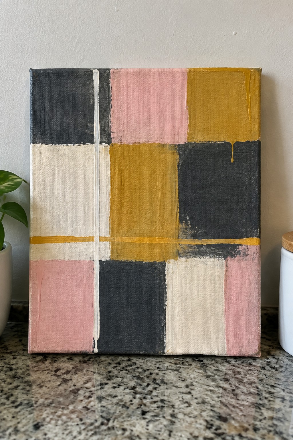 An abstract painting on canvas with a grid of black, pink, yellow, cream, and gray rectangles plus paint drips, sitting on a granite countertop.