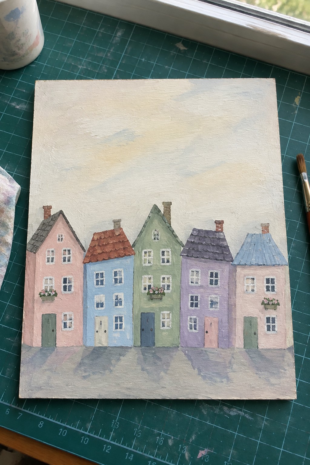 Colorful row houses painting on canvas atop green gridded cutting mat with paintbrush nearby
