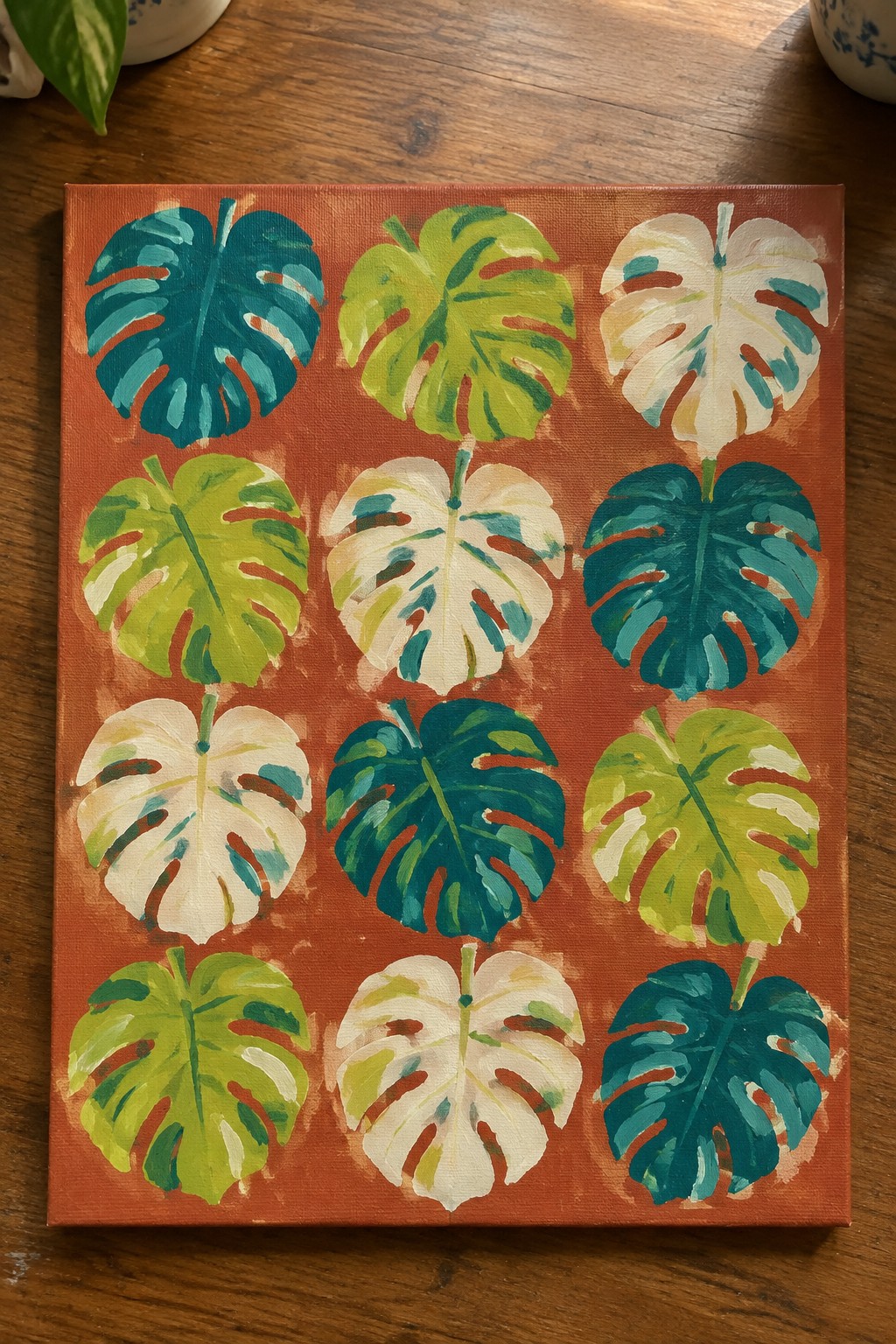 Vibrant monstera leaves in teal, green, and white on orange canvas on wood
