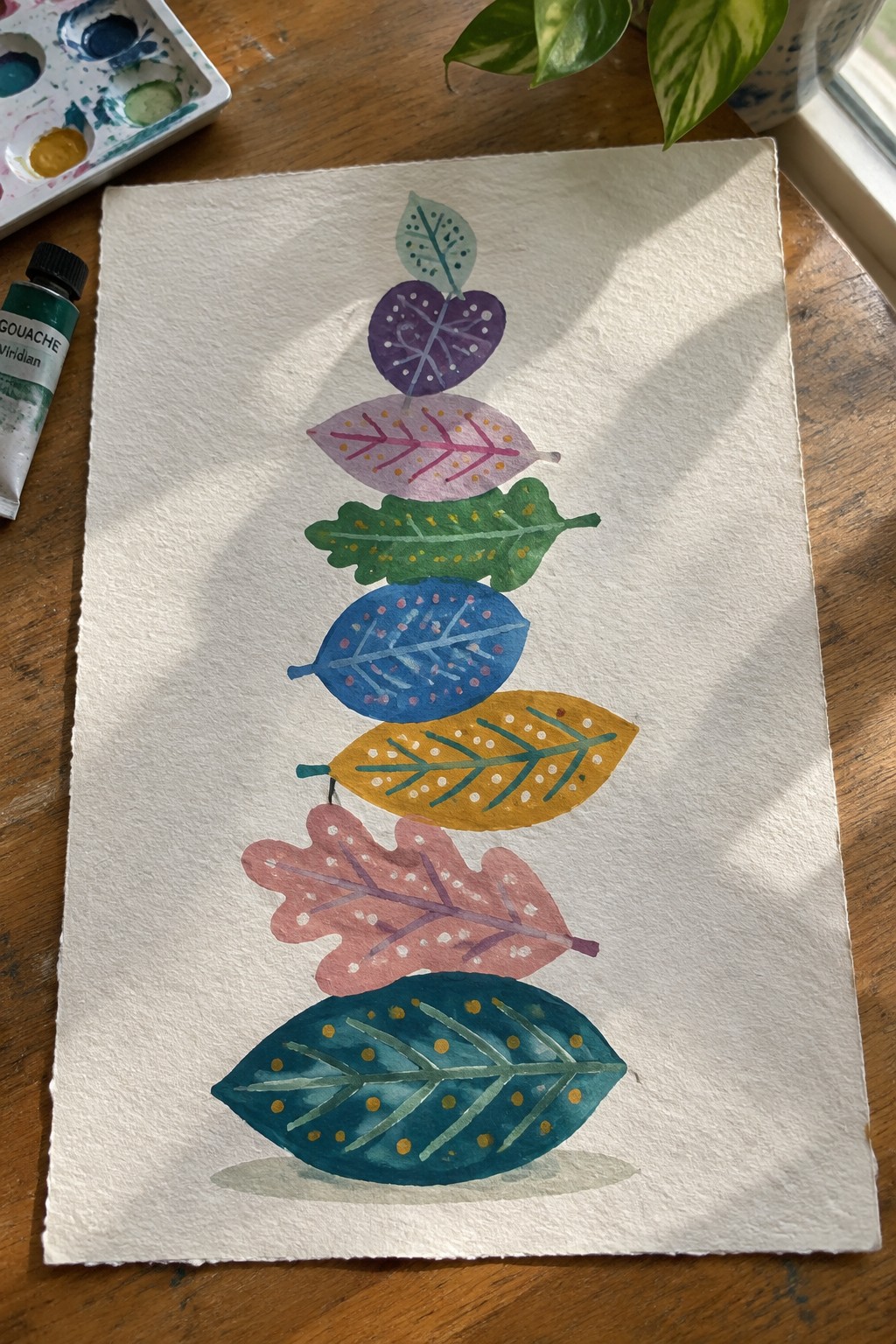 Colorful stacked leaves painted in watercolor on white paper with gouache paints nearby