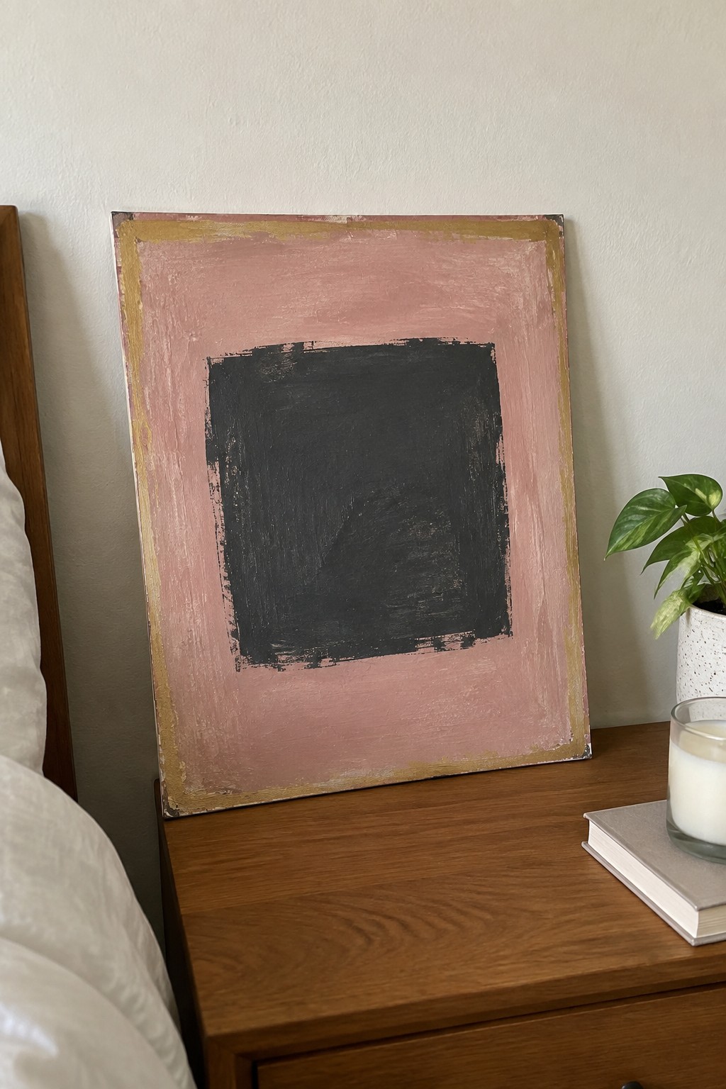 Abstract pink painting with black square on wooden nightstand beside plant and candle