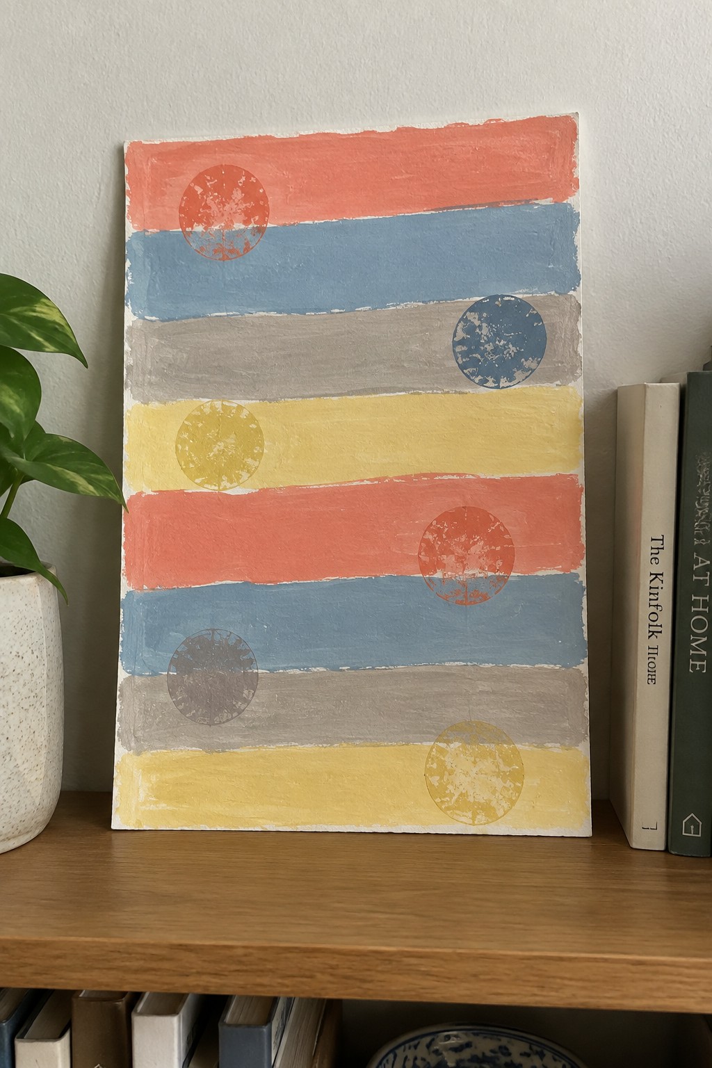 Colorful abstract painting with horizontal stripes and circles on shelf beside books and plant