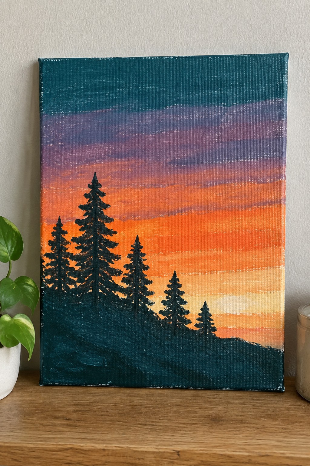 A rectangular canvas painting shows a sunset sky with horizontal bands of teal, purple, orange, and yellow above dark pine tree silhouettes on a sloped hill.