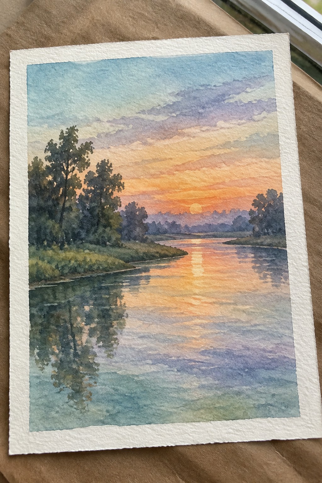 Watercolor sunset over calm river with orange sun reflection, trees, and colorful sky.